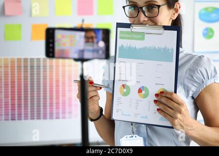 Woman in office holds charts in her hand and demonstrates them through a smartphone Stock Photo