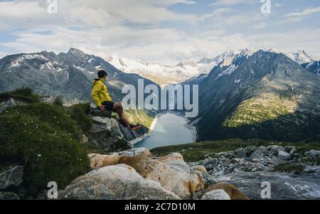 Adventurous man on the edge of a cliff overlooking the beautiful Austria Alps and Lake during a vibrant summer sunset. Taken in Hiking to the Olperer Stock Photo