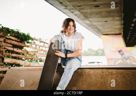 Teenage girl in stylish clothes posing on half pipe ramp an outdoor skate park. Beautiful kid female model skateboarder with skate board in urban Stock Photo