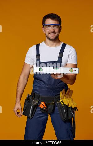 Young caucasian repairman worker in safety glasses and uniform showing builder level. handyman wearing tool belt isolated on orange studio background Stock Photo