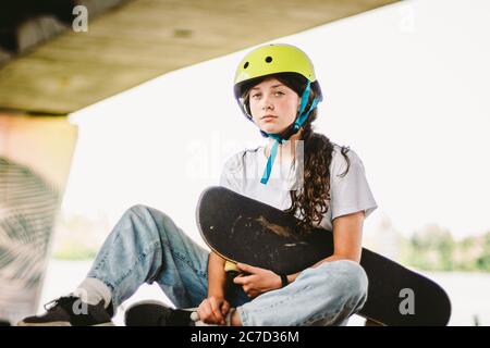 Teenage girl in helmet and stylish clothes posing on half pipe ramp an outdoor skate park. Beautiful kid female model skateboarder with skate board in Stock Photo