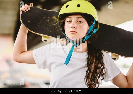 Teenage girl in helmet and stylish clothes posing on half pipe ramp an outdoor skate park. Beautiful kid female model skateboarder with skate board in Stock Photo