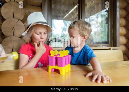 Two cute adorable caucasian blond little sibling sitting at table home yard and have fun tasting mom homemade fruit icecream. Young happy sister and Stock Photo