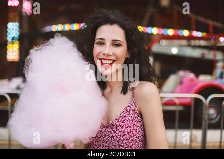 Portrait of smiling lady with dark curly hair in dress standing with cotton candy in hand and happily looking in camera while spending time in Stock Photo