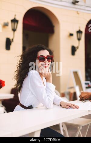 Beautiful smiling lady with dark curly hair in sunglasses standing and happily looking in camera while spending time in summer terrace of cafe Stock Photo