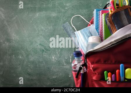 Backpack full of school supplies with mask and gel protection for covid-19 with green blackboard with blank space for writing front view Stock Photo