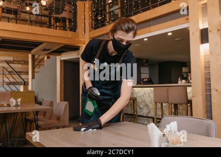 A waitress who wears a black medical face mask and disposable medical gloves is holding a bottle with sanitizer and cleaning a table with a rag in a r Stock Photo