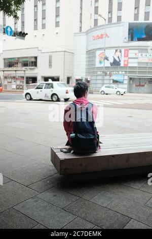 MATSUYAMA, JAPAN - Sep 21, 2019: A vertical selective shot of a person wearing black backpack sitting on a bench  on the sidewalk near cars and buildi Stock Photo