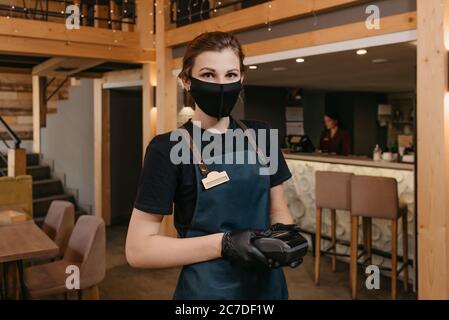 A waitress who wears an apron, a black medical face mask, and disposable medical gloves is holding a wireless payment terminal in a restaurant. The st