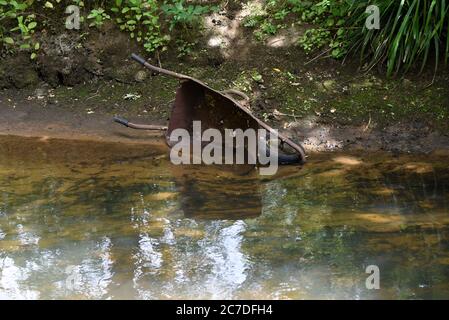 A rusty wheelbarrow reflected in water as it ends its days in the Blackwater River on the border between Hampshire and Berkshire Stock Photo