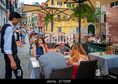 Man with a gondola passes an outdoor restaurant terrace by a canal in Venice, Italy. Tourists dining at the popular outdoor Ristorante in Fondamenta d Stock Photo