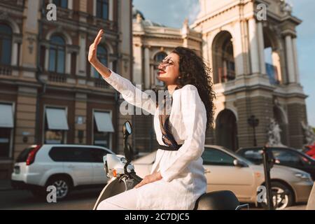 Pretty smiling woman with dark curly hair in white costume sitting on moped and happily covering her face with hand from sunlights with city view on Stock Photo