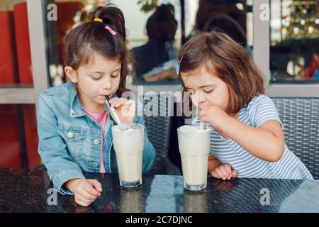 Two funny Caucasian preschool sisters siblings drink sip milk shakes in cafe. Friends girls eating brunch breakfast outdoor. Cold summer desserts for Stock Photo