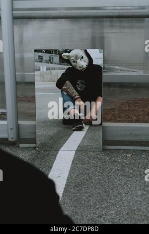 black hooded person with a sheep mask staring at his reflection in the mirror on a pavement Stock Photo