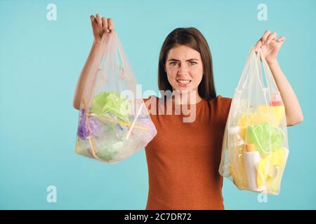 Emotional girl squeamishly holding eco bags with plastic waste displeasingly looking in camera over colorful background Stock Photo