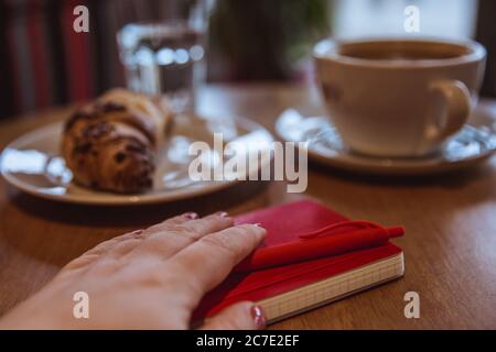 A red note and red pen is on the table in a cafe, a cup of coffee and a croissant on background. Female hand holding a notepad Stock Photo