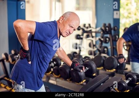 A portrait of bald senior man feeling strong back pain during training in the gym. People, healthcare and lifestyle concept Stock Photo