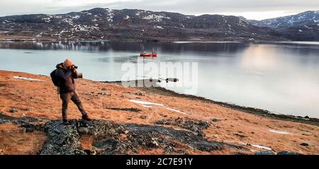 Photographer taking photo whilst stood on rock in Greenland with boat and fjord in the background Stock Photo