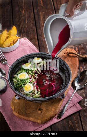 Hand pours from a white jug ?old beetroot soup with eggs. Baked potatoes, souce, pink tablecloth on the rustic wooden background. Top view Stock Photo
