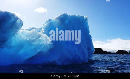 Glacial blue iceberg floating in dark antarctic ocean water under blue sky with dark rocks in background Stock Photo