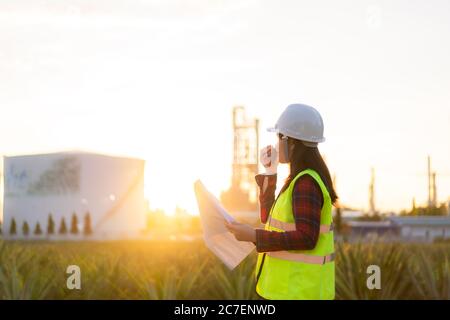 Asian woman technician Industrial engineer using walkie-talkie and holding bluprint working in oil refinery for building site survey in civil engineer Stock Photo