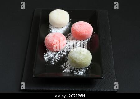 Four types of Japanese dessert mochi - pomegranate with honey, green matcha tea, strawberry, coconut on a black plate on a black table. Closeup Stock Photo