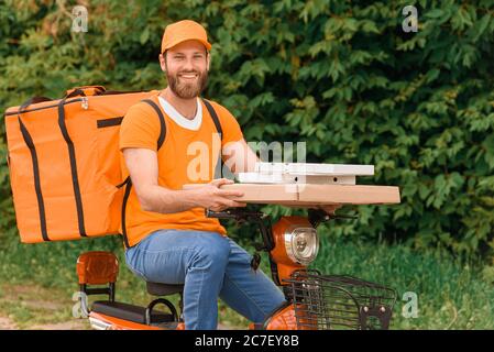 Food delivery man in orange uniform sitting on a moped with a food delivery bag and holding boxes of pizza. Stock Photo