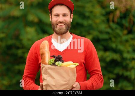Portrait of a food delivery man in a red uniform holding a paper eco bag with groceries in his hands. Stock Photo