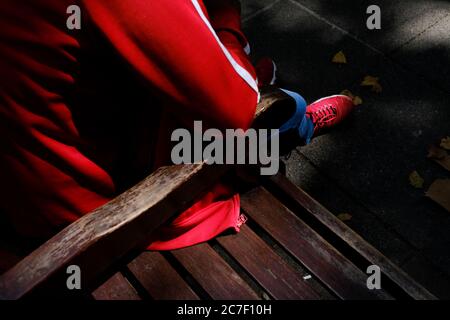High angle shot of a person with a red jacket and sneakers sitting on a wooden bench during daytime Stock Photo