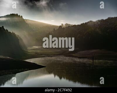 Beautiful shot of a pond surrounded by forested mountains with the sun shining behind the clouds Stock Photo
