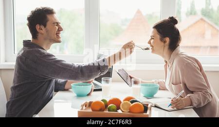 Side view photo of a caucasian couple feeding each other while using a laptop with tablet and eating fruits Stock Photo