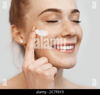 Freckled caucasian ginger lady applying on face anti aging cream and smile on a white studio wall Stock Photo