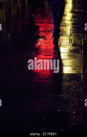 Vertical shot of people walking on the wet pavement reflecting the lights at night Stock Photo
