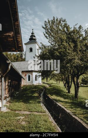 Old white church with tower on the edge of the old Slovenian village with the stream flowing around and shadows below the trees in garden Stock Photo