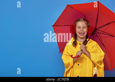 Happy funny child with red umbrella posing on blue wall background. Girl is wearing yellow raincoat and rubber boots. Stock Photo