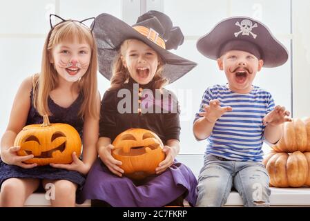 Happy brother and two sisters on Halloween. Funny kids in carnival costumes indoors. Cheerful children play with pumpkins. Stock Photo