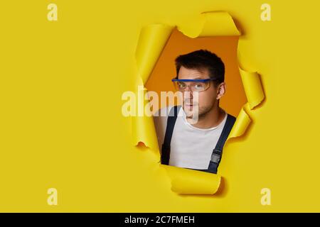 caucasian handyman worker in safety glasses and uniform looking through hole in yellow paper. copy space Stock Photo