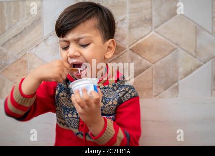 Cute little boy in red dress, eating cup ice cream with spoon at home. Stock Photo