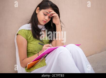 Indian late teen girl student sitting near wall and doing study. She is reading and writing on book in outdoor at day time. Stock Photo
