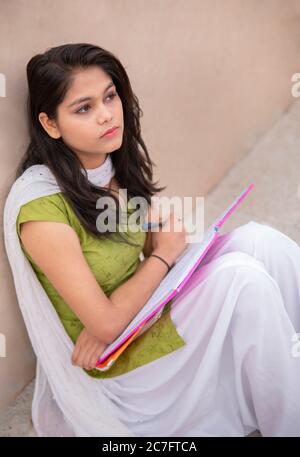Indian late teen girl student sitting near wall, doing study and thinking while reading on book in outdoor at day time. Stock Photo