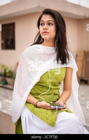 Beautiful, happy Indian late teen girl listening music on smartphone through earphones and enjoying fresh air in outdoor on roof at day time. Stock Photo