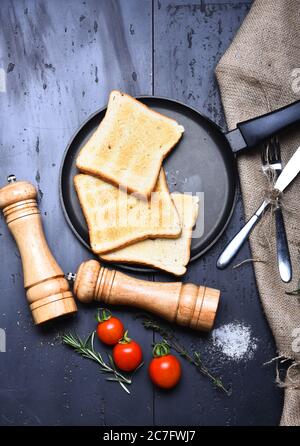 Slices of roast bread with tomatoes, thyme, fork and knife. Breakfast at home, restaurant. American tradition concept. Toasts on frying pan on grey background near sackcloth, saltcellar and pepperbox Stock Photo