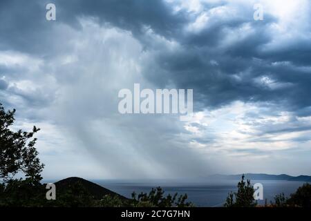 A wall of rain falls from a dark cloud over the sea and the storm seems to approach. Stock Photo