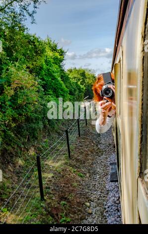 Candid view of a woman photographer, leaning out of a vintage passenger train while in motion, using a camera to photograph the steam locomotive. Stock Photo