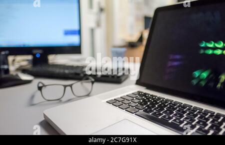 Close-up, shallow focus view of a well-known computer laptop on an office desk together with a server connected monitor and a pair of glasses. Stock Photo