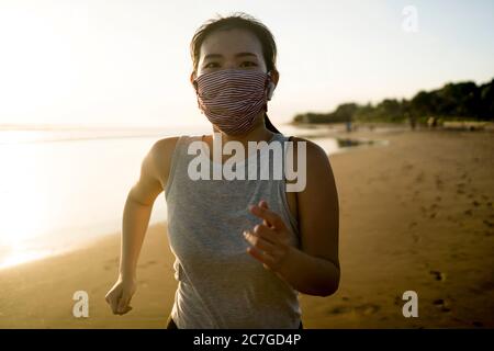 new normal running workout of Asian girl in face mask - young happy and attractive Chinese woman jogging on the beach in post quarantine outdoors exer