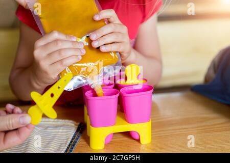 Cute adorable young caucasian little girl kid sitting at table home yard and have fun making with mom homemade fruit puree icecream. Young happy child Stock Photo