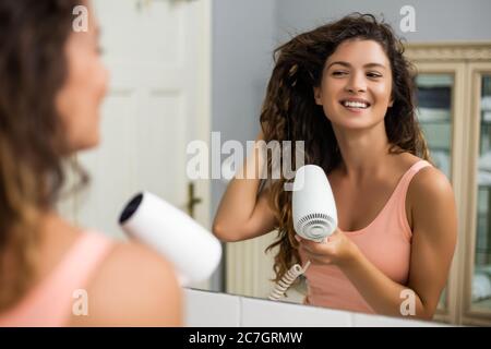 Young woman with gorgeous curly hair using hairdryer. Stock Photo