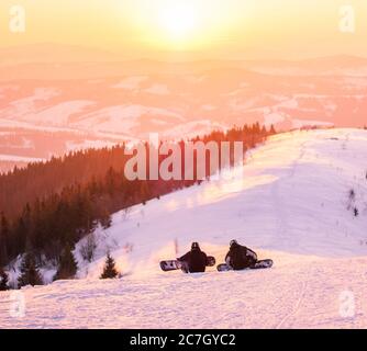 High angle shot of two skiers sitting on the bright snow watching the sunset with forest in distance Stock Photo