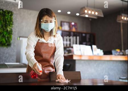 New normal startup small business Portrait of Asian woman barista wearing protection mask and face shield cleaning table in coffee shop while social d Stock Photo